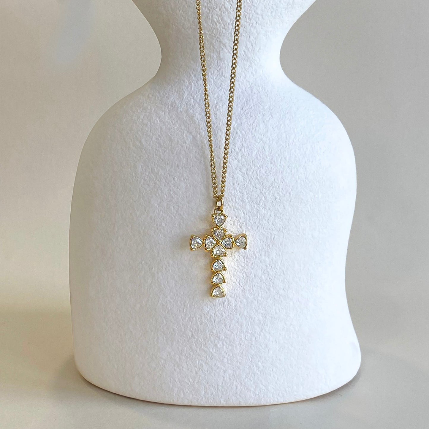 Colgante de cruz de Bendita  Eva en acero y oro mostrado en busto. Circonitas en forma de corazón, 28x19 mm. Diseño elegante y moderno, perfecto para looks de tendencia.